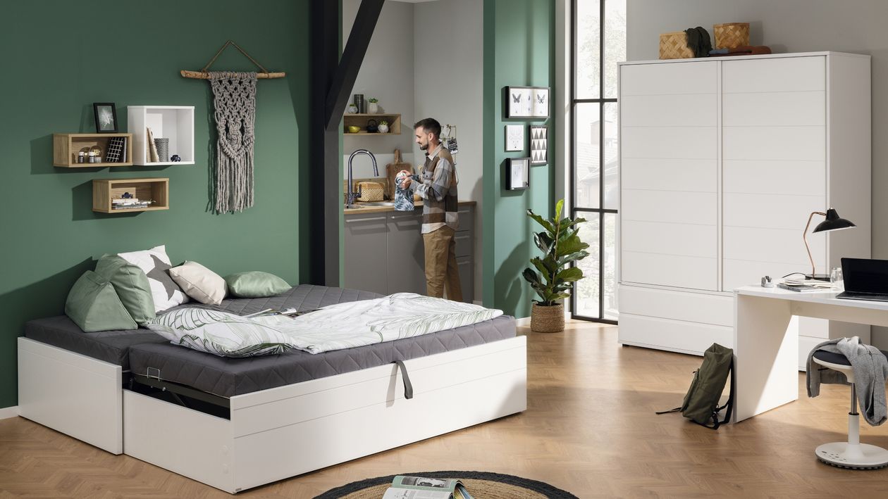 Das Funktionsbett Flynn von Paidi steht in einem kleinen Zimmer mit integrierter Küche und Homeoffice. An der Spüle trocknet ein Mann Geschirr.