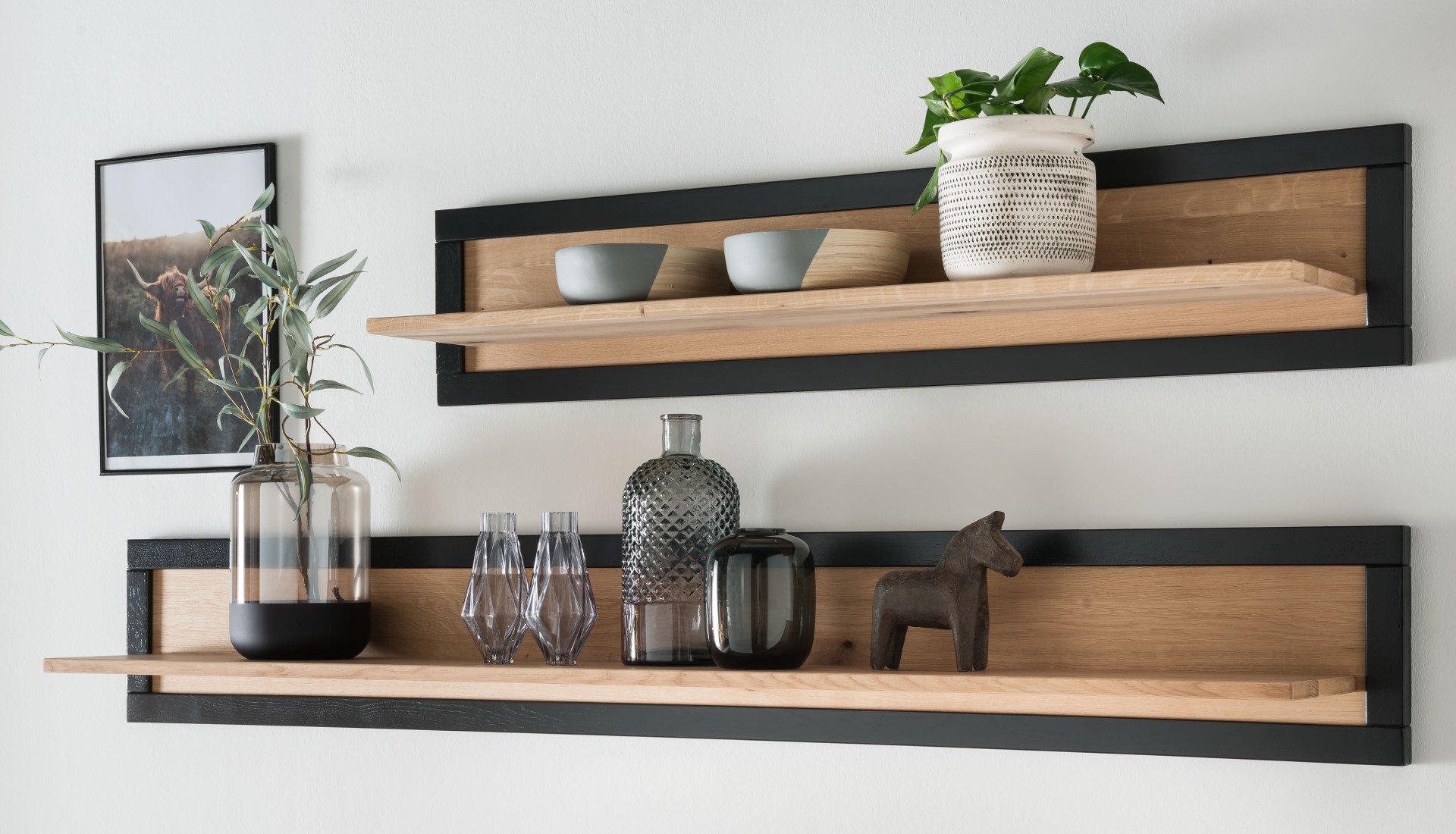 Zwei Holzregale mit schwarzen Rahmen halten Vasen, Schalen, eine Topfpflanze und ein kleines Holzpferd an einer hellen Wand.