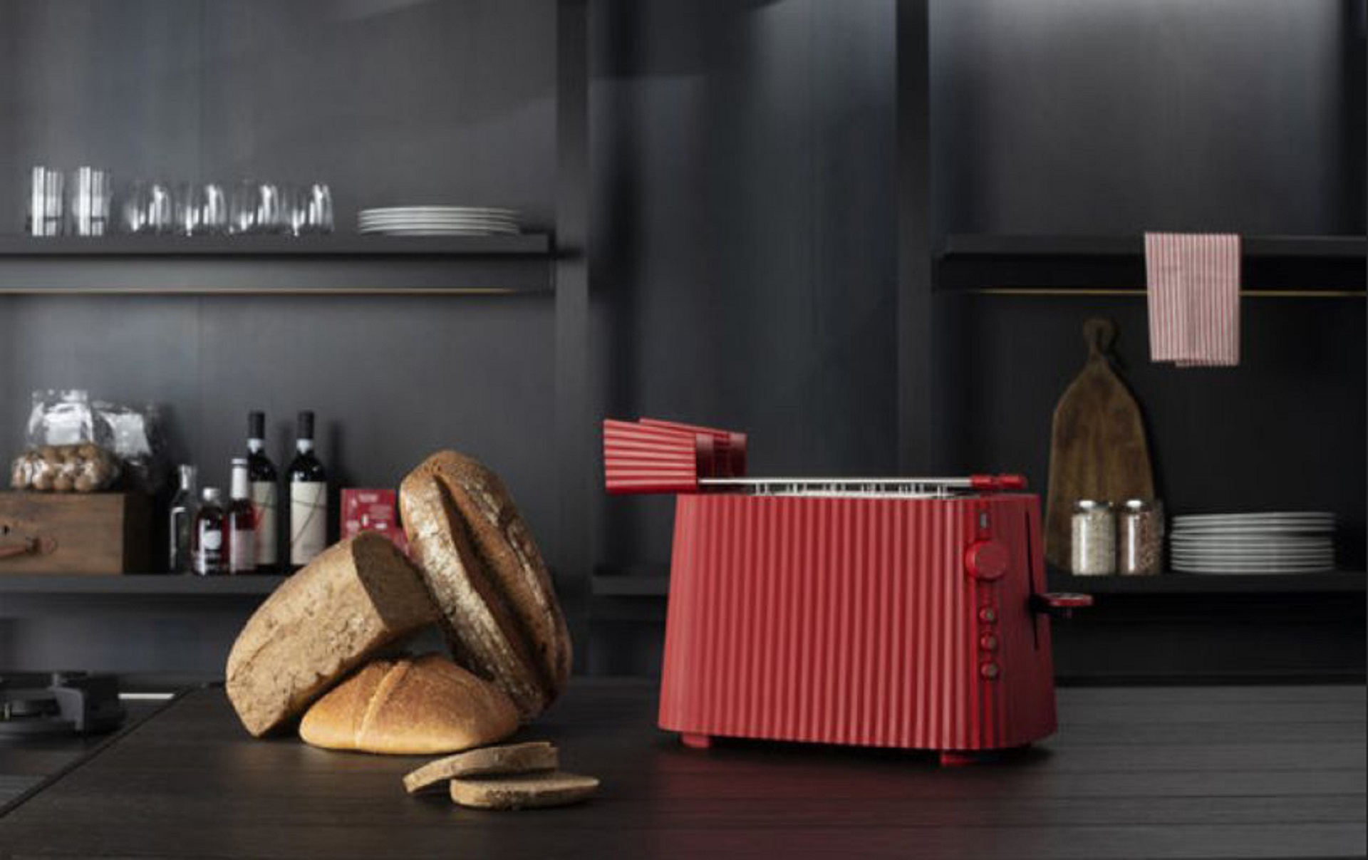 Toaster Plisse aus dem Hause Alessi aus rotem Kunststoff auf einem modernen, dunklen Küchentresen neben Brot und weiteren Küchenutensilien