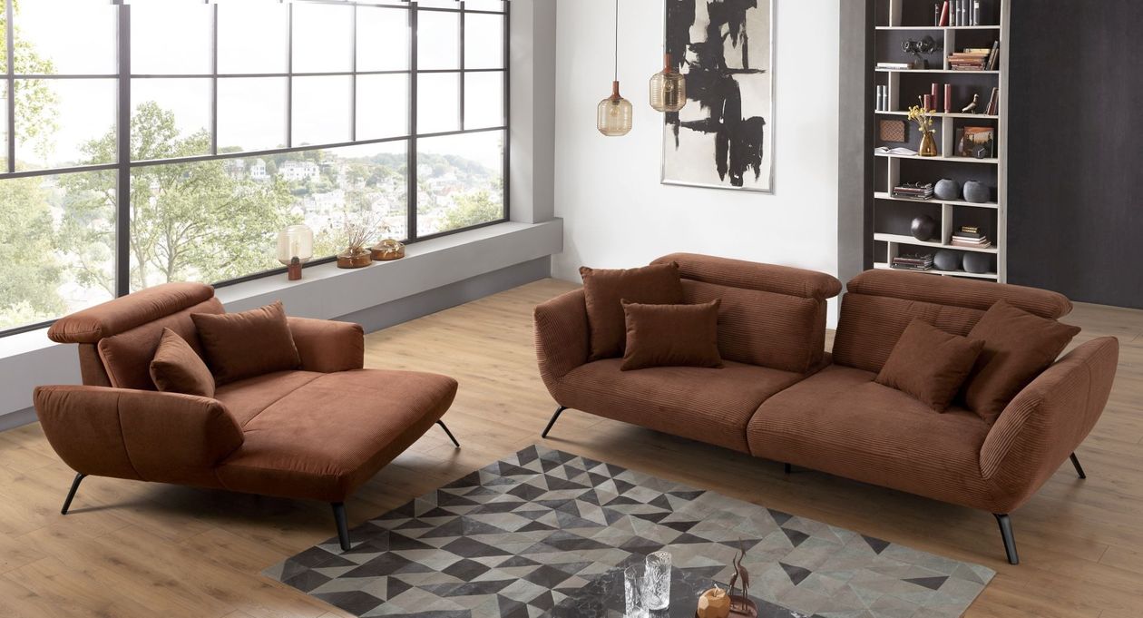 Rechts befindet sich das kupferfarbene Megasofa "Seattle" aus Cord im Retro-Stil in Kombination mit einem passenden Sessel links daneben. Der Raum ist im modernen Industial-Stil mit einem großen Fenster eingerichtet und im Hintergrund befindet sich ein weißes Holz-Bücherregal..