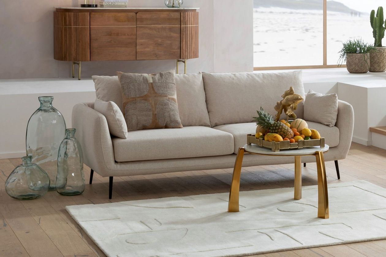 Auf dem Foto befindet sich ein beiges 2-Sitzer Sofa mit dunklen Füßen aus Metall. Davor befinden sich ein weißer Teppich und ein weißer Couchtisch mit goldenen Beinen und einer Obstschale.