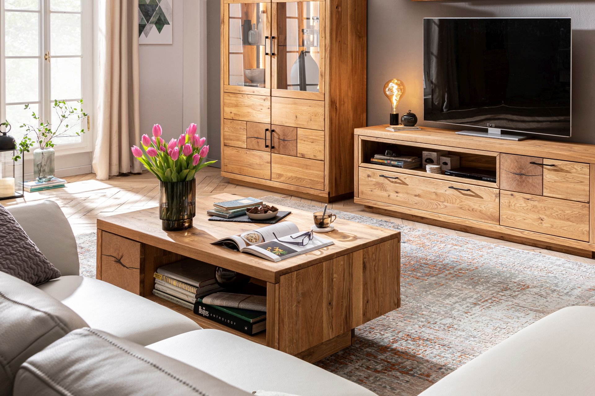 Im Bildausschnitt befindet sich ein massiver Couchtisch aus geöltem Eichenholz vor einem beigen Sofa. Im Hintergrund befinden sich das dazu passende Highboard und Lowboard mit einem Fernseher.