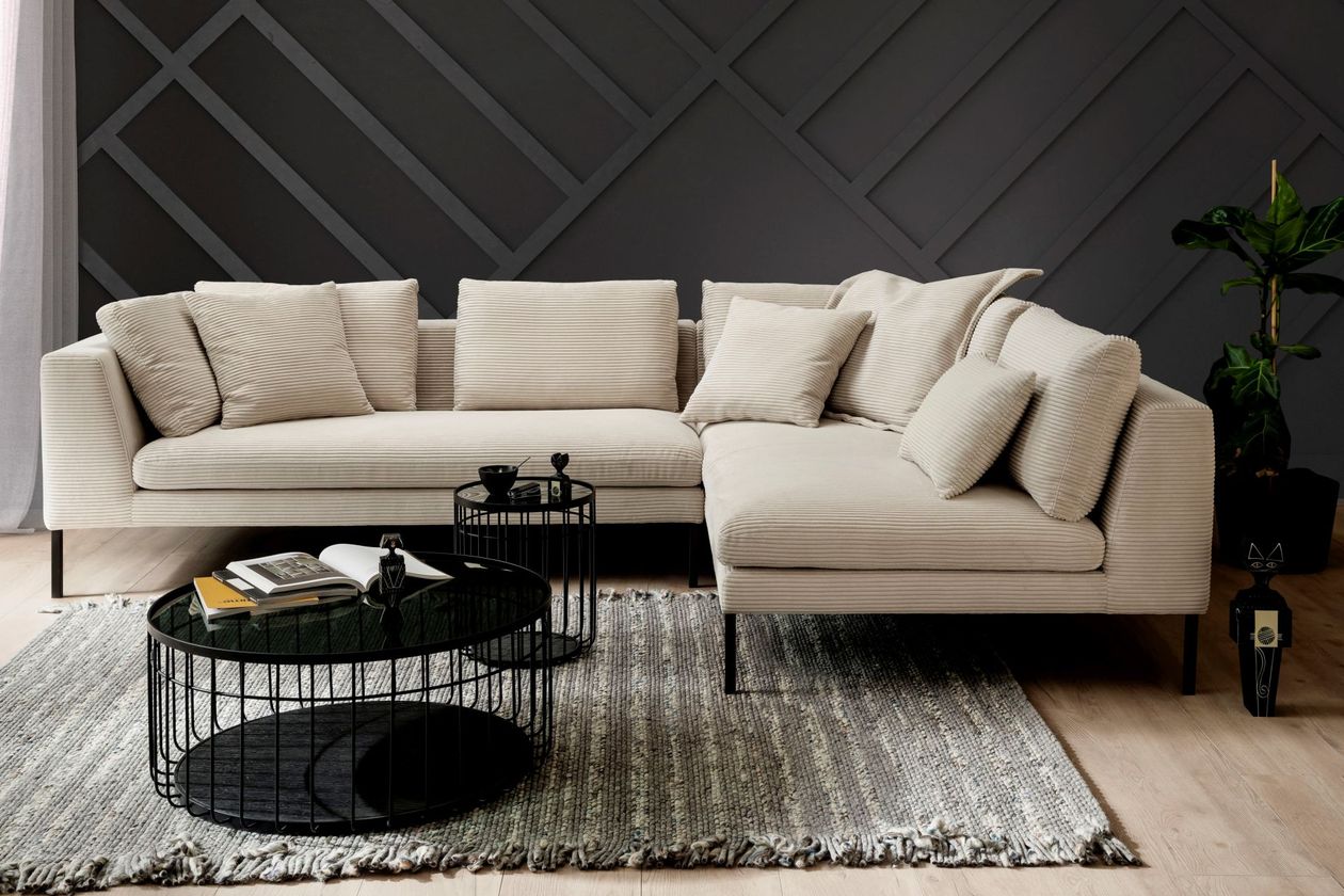 Auf dem Foto befindet sich ein beiges Cord-Ecksofa mit schwarzen Metallbeinen umgeben von schwarzen Wänden. Unter dem Sofa liegt ein hellgrauer Teppich, auf dem zwei schwarze Couchtische mit dunkler Glasplatte und Metallgitter-Optik stehen.