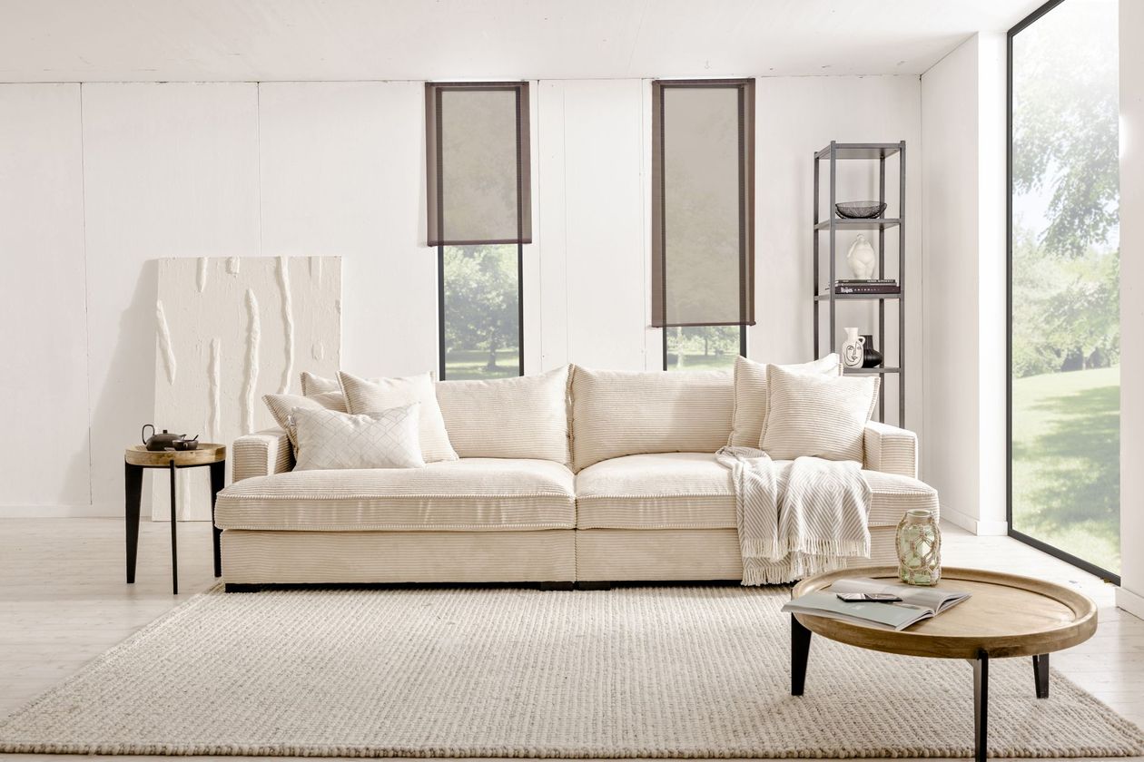 In der Mitte des Bildes befindet sich das cremefarbene Bigsofa "Tribecca" aus Cord von BOOOM. Der Raum ist hell und in cremefarbenen Tönen gehalten mit Akzenten in Grau-schwarz.