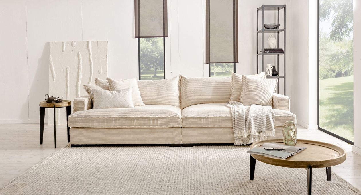 In der Mitte des Bildes befindet sich das cremefarbene Bigsofa "Tribecca" aus Cord von BOOOM. Der Raum ist hell und in cremefarbenen Tönen gehalten mit Akzenten in Grau-schwarz.