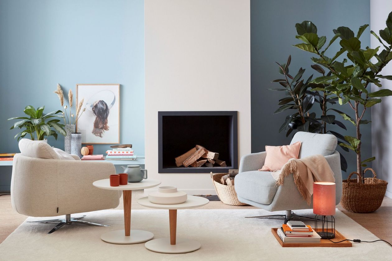 Auf dem Foto befinden sich zwei beige Drehsessel vor einem hellen Kamin. Darunter befindet sich ein weißer Teppich mit einem 2-er Satz weißer Beistelltische.