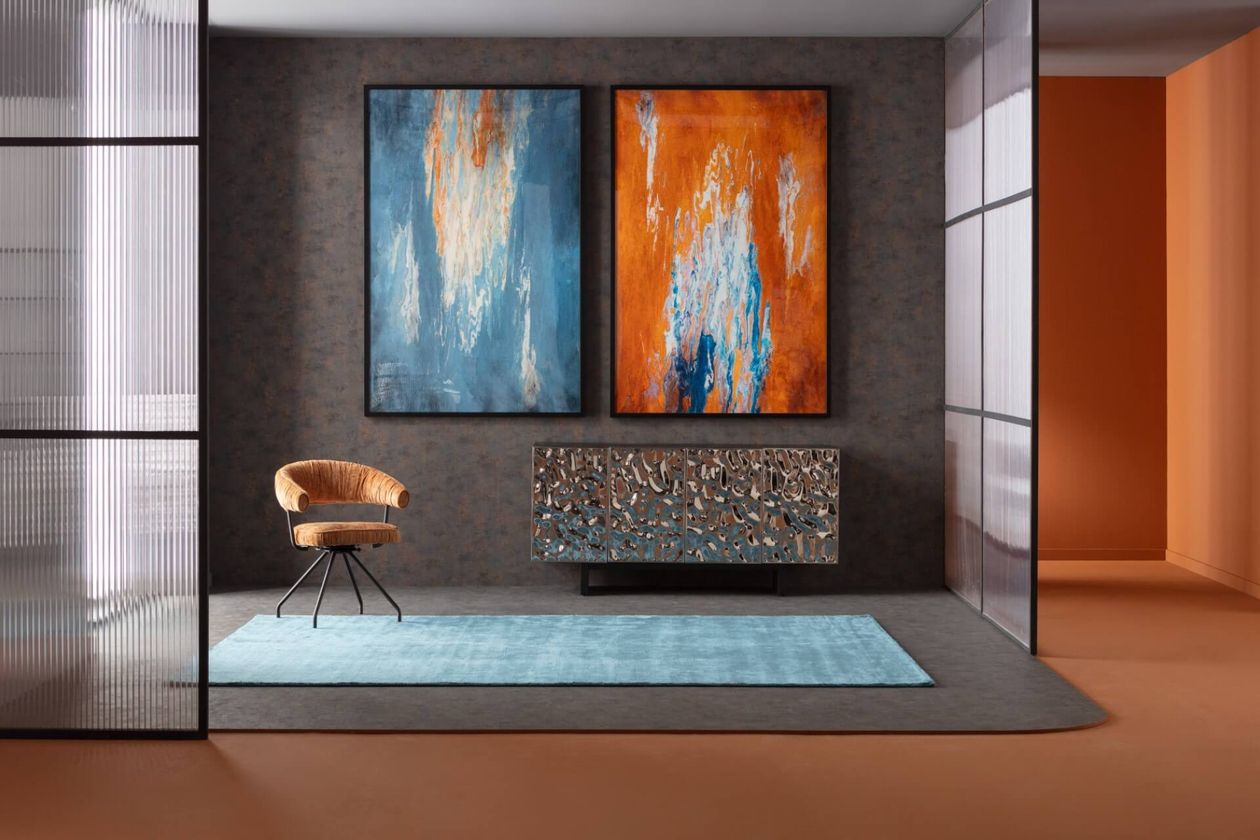 Modernes Zimmer mit abstrakten blau- und orangefarbenen Gemälden, mit verschiedenen Möbeln der Marke KARE Design, wie einem strukturierten Sideboard, einem beigen Stuhl und einem blauen Teppich auf einem grauen Boden.