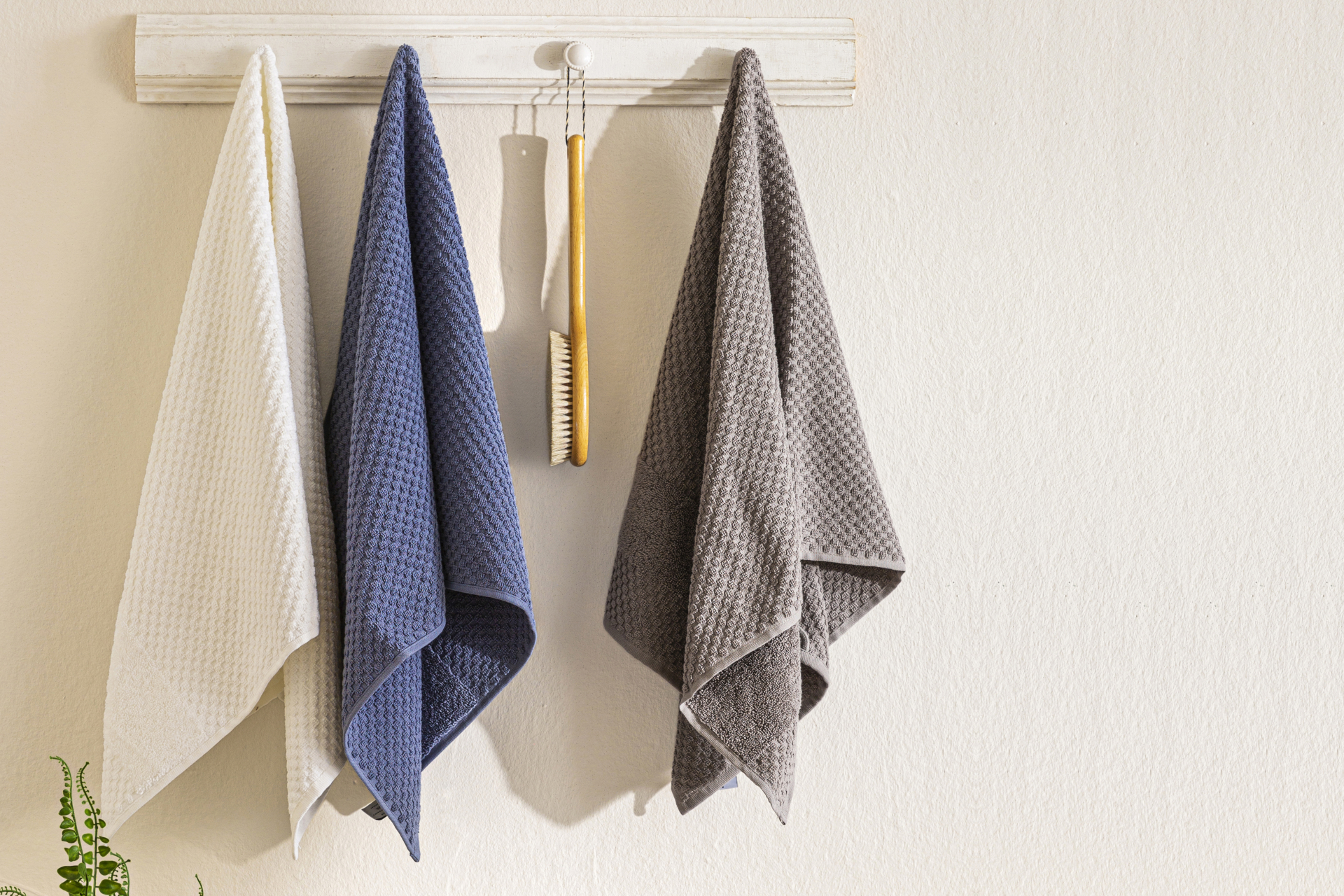 Mehrere Baumwollduschtücher der Serie Isabelle von Fürstenhof hängen an einer weißen Hakenleiste. Dazwischen hängt eine hölzerne Duschbürste.
