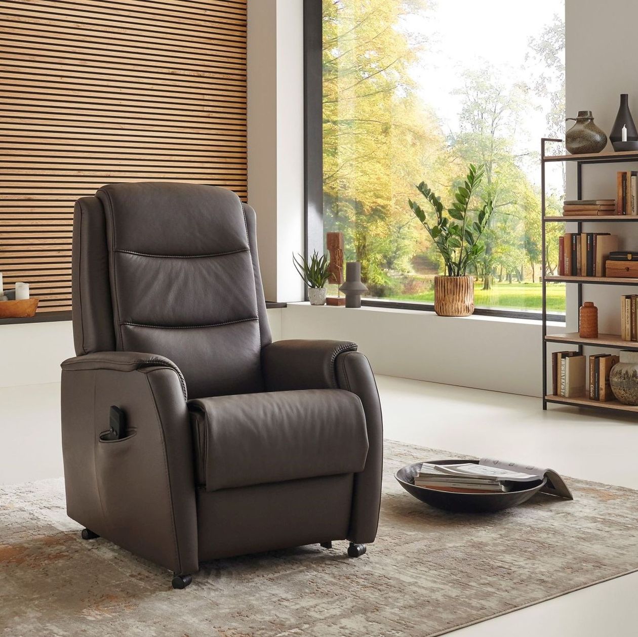 Ein dunkelbrauner Relaxsessel aus Leder auf einem beige-braunen Teppich in einem hellen Raum mit vielen Pflanzen und einem Regal im Hintergrund.