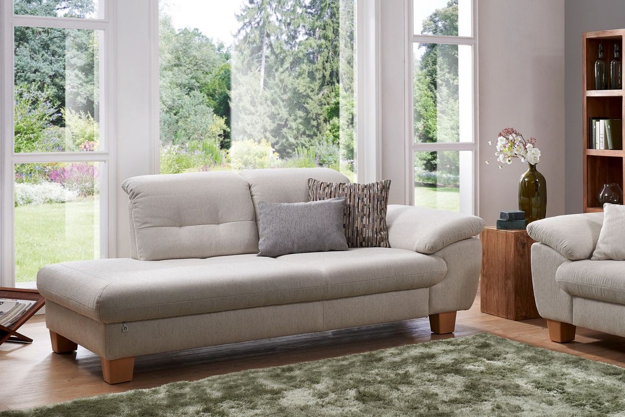 Auf dem Foto befindet sich eine cremefarbene Recamiere mit Stoffbezug und hölzernen Füßen. Im Hintergrund ist ein großes Fenster, welches zu einem Garten hinaus zeigt und vor dem Sofa liegt ein grüner Teppich.