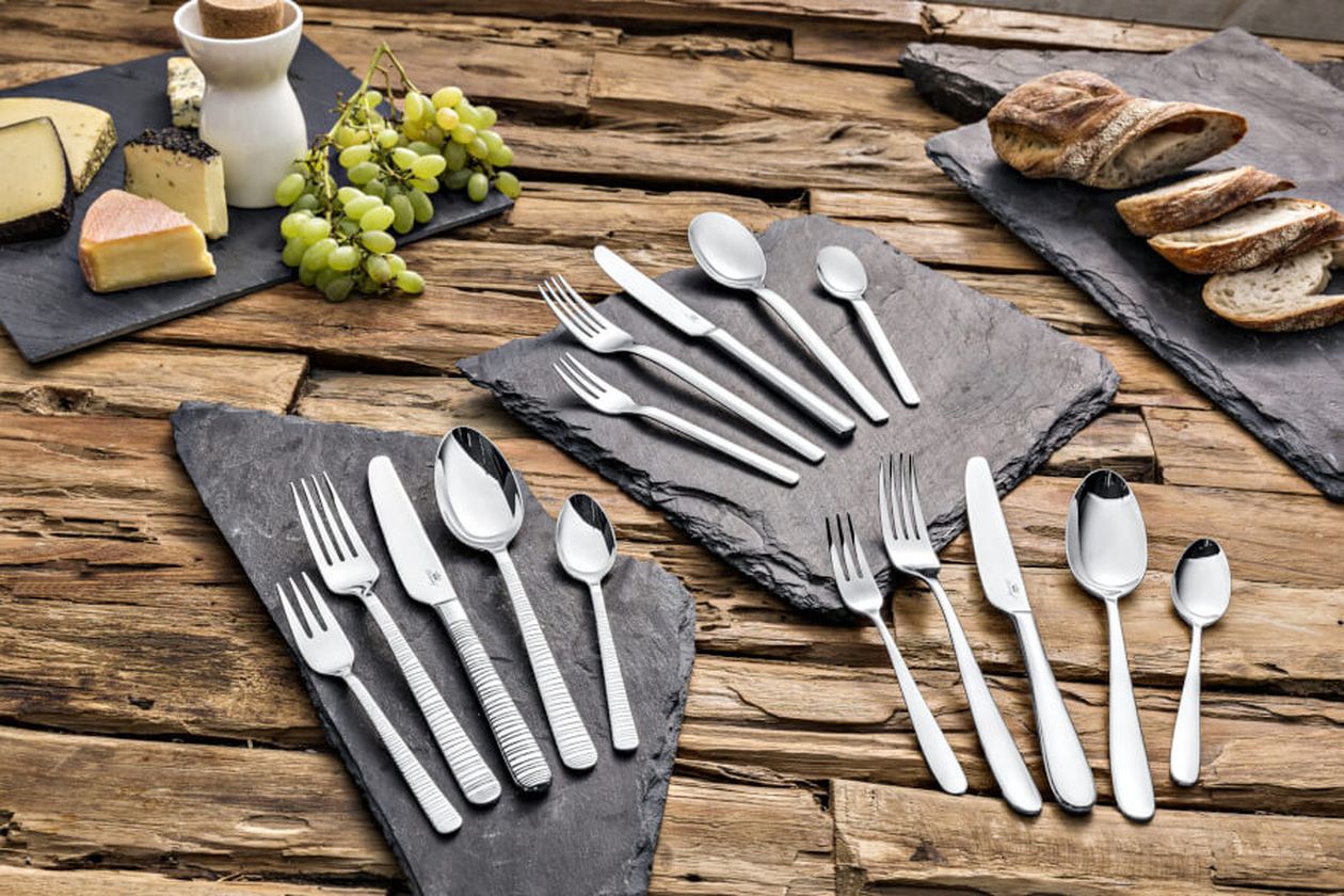 Die drei silbernen Edelstahl-Bestecksets aus dem Hause Fürstenhof Amalia, Charlotte und Ariane auf einer rustikalen Echtholzplatte mit Platzsets aus Schiefer.