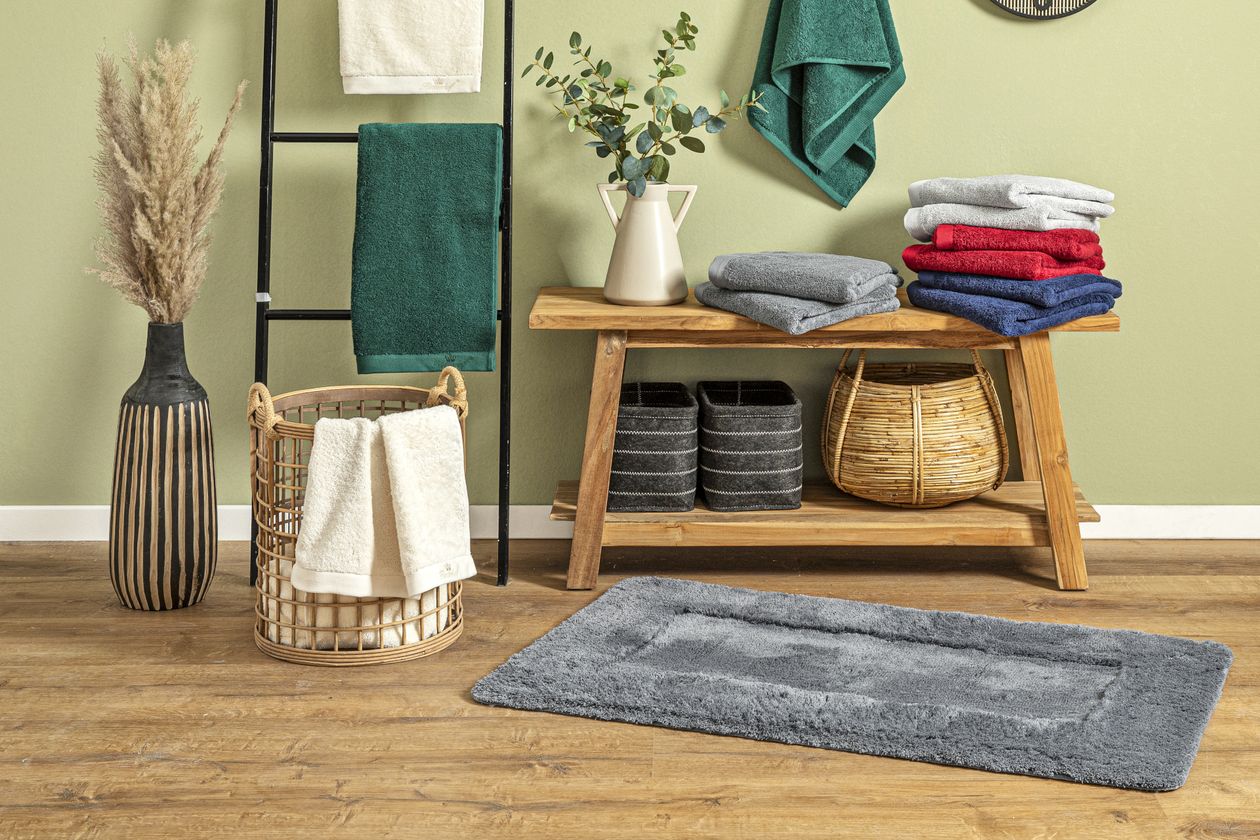 Der graue, aus Polyacryl gefertigte Badteppich Viktoria von Fürstenhof vor einer Holzbank mit Handtüchern und verschiedenenKörben