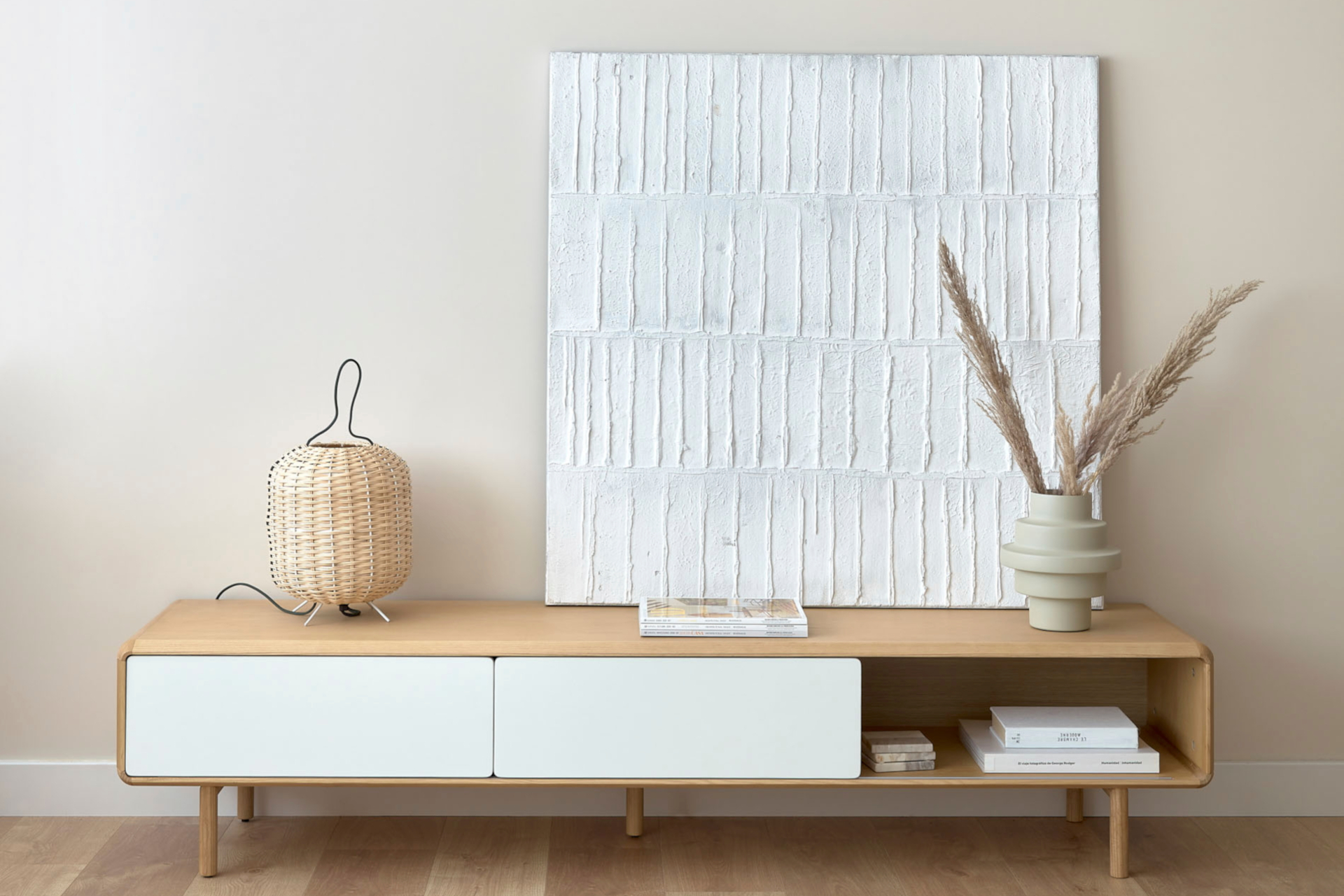 Einrichten mit Kave Home: Das Wandbild Basilsa aus weiß bemaltem Segeltuch steht auf dem natürlich dekorierten TV-Lowboard Anielle aus Holz mit weißen Schiebetüren.