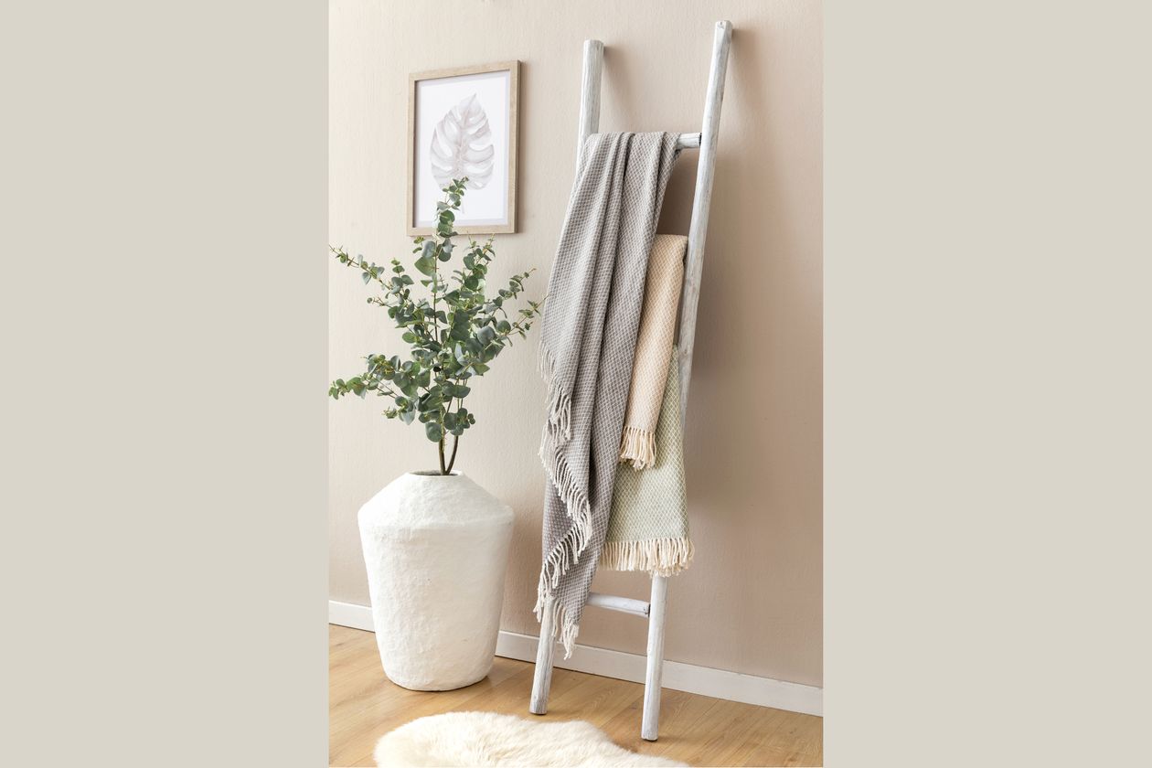 Drei Modelle des Plaids Amalia von Fürstenhof mit Fransen und in den Farben Grau, Natur und Salbei hängen übe eine weiße Leiter. Daneben steht ein großes, weißes Pflanzgefäß mit einem kleinen Bäumchen.