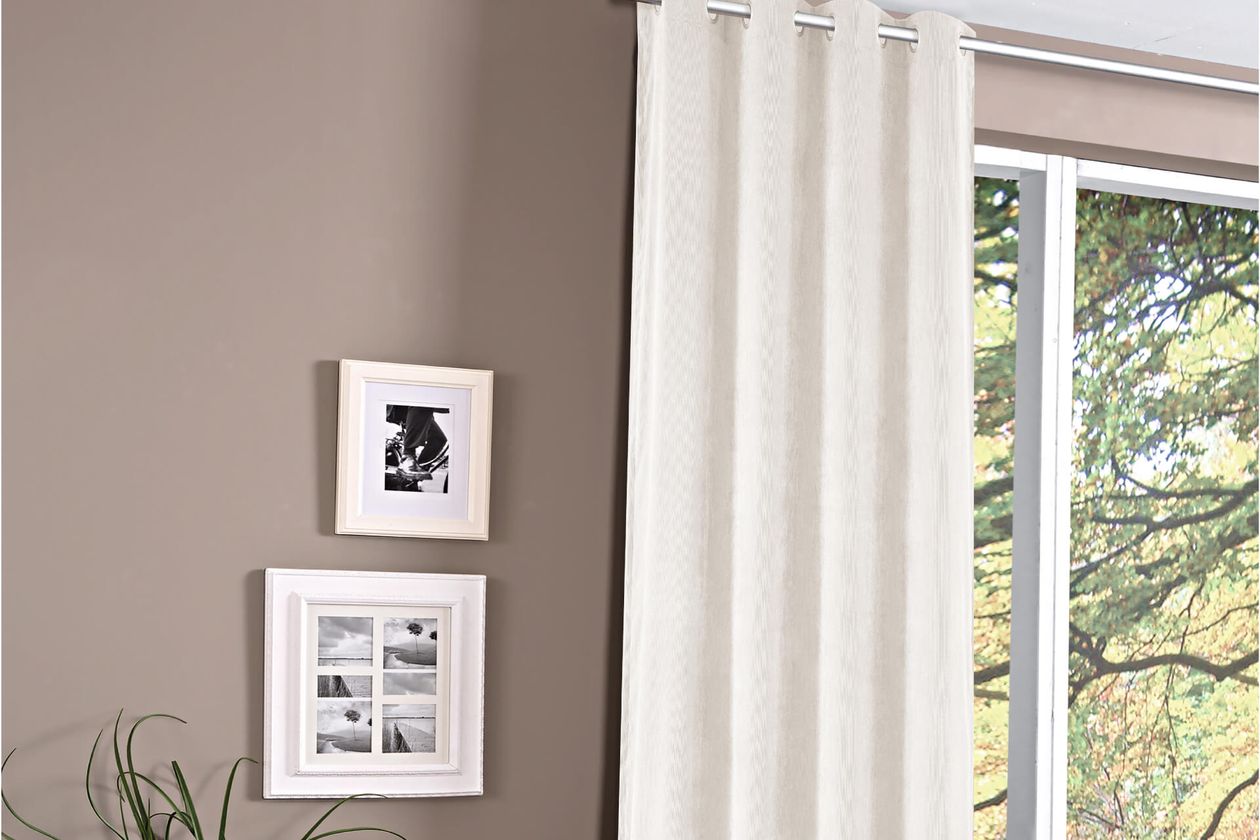 Weiße Vorhänge hängen neben einem großen Fenster mit Blick auf Bäume. An der angrenzenden beigen Wand hängen zwei gerahmte Schwarz-Weiß-Fotos.