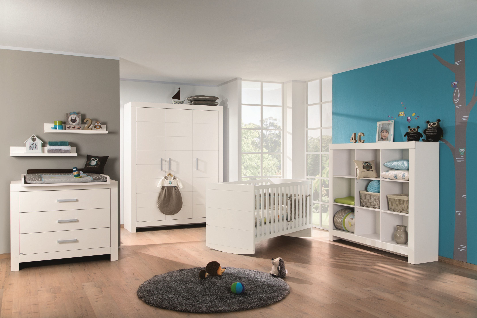 Ein verspieltes Babyzimmer eingerichtet mit den weißen Möbeln der Paidi Serie Fiona. Eine Wand ist blau gestrichen und mit der Zeichnung eines Baumes verziert.