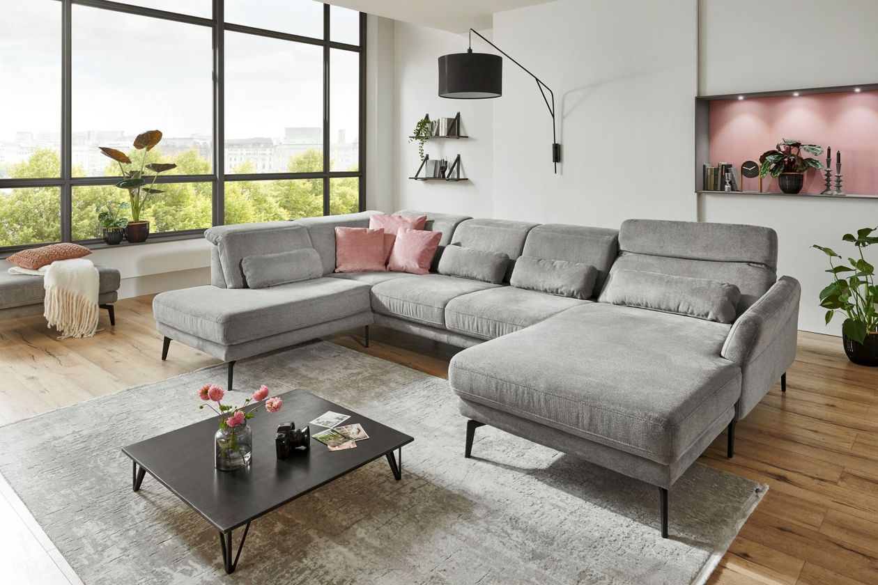 Auf dem Foto befindet sich eine U-förmige hellgraue Wohnlandschaft mit schwarzen Beinen und ausklappbaren Kopfstützen. Darunter befindet sich ein grauer Teppich auf dem ein schwarzer Couchtisch steht. Der Raum ist hell eingerichtet mit Dekohighlights in Rosa.