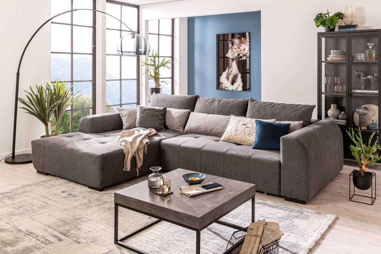Auf dem Foto befindet sich ein dunkelgraues Ecksofa auf einem hellgrauen Teppich mit einem dunkelgrauen Couchtisch. Im Raum sind Highlights mit blauer Deko und einer blauen Akzentwand gesetzt.