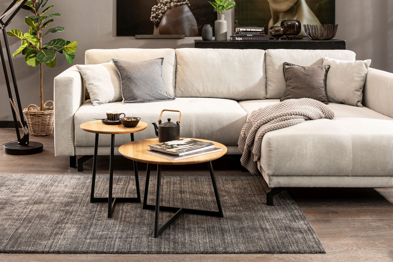 Im Fokus des Bildes befindet sich zwei runde Couchtische mit Holzplatte und schwarzen Metallbeinen auf einem dunkelgrauen Teppich. Dahinter steht ein helles Cord-Ecksofa. Der Raum ist, bis auf das Sofa, in schwarzen und dunkelgrauen Tönen gehalten.