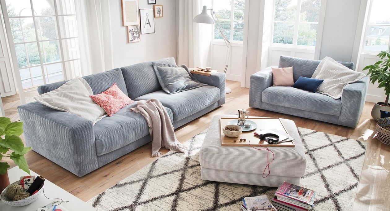 Ein hellblaues Cord-Sofa und ein hellblauer Cord-Sessel ausgerichtet in einer L-Form. Der helle Raum ist mit weißen Vorhängen, einer weißen Scandi-Stehlampe, einem weißen Cord-Hocker als Couchtisch-Ersatz und einem weißen Teppich mit einem dunklen Rautenmuster ausgestattet.