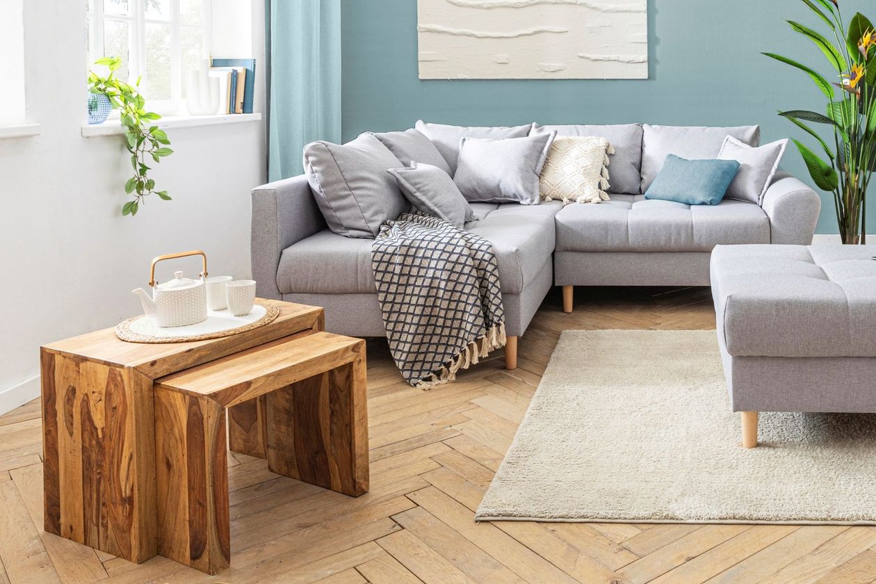 Vorne rechts befindet sich ein Couchtisch-Set bestehend aus zwei Tischen in umgedrehter U-Form, die halb übereinander geschoben sind. Links im Hintergrund steh ein graues Ecksofa im skandinavischen Stil mit einem passenden Fußhocker.