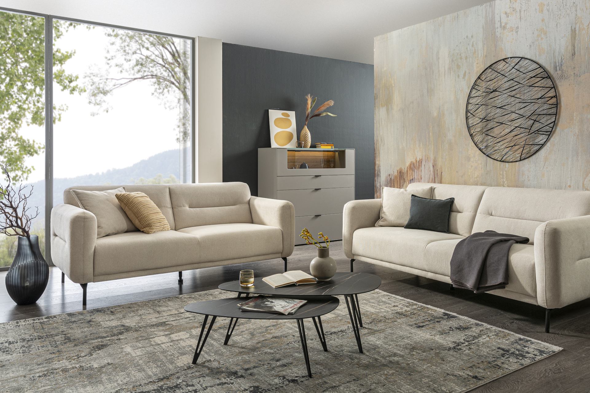 In einem stilvollen Wohnzimmer stehen vor einer großen Fensterfront und um ein Set aus filigranenn schwarzen Couchtischen zwei Sofas des Typs Nova der Marke BOOOM in Beige mit schwarzen Füßen.