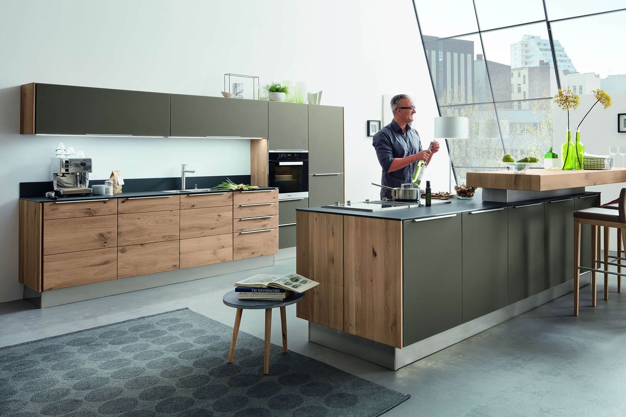 Moderne Küche mit Holz und dunklen Schränken, Kochinsel und einem Mann, der Essen zubereitet. Große Fenster bieten einen Blick auf die Stadt. Zeitgenössische Einrichtung.