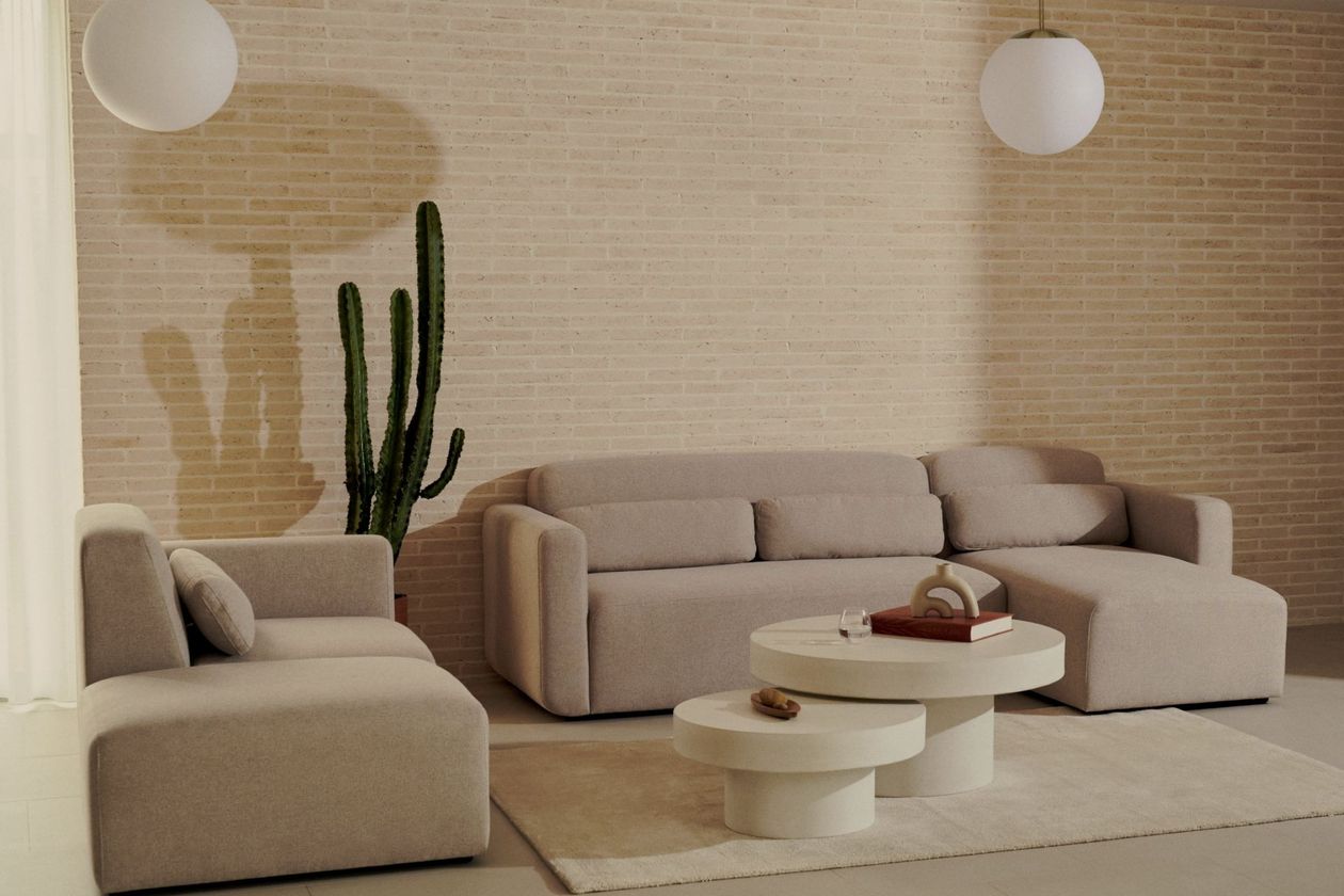 Hinten an einer hellen Ziegelwand steht ein beiges Sofaelement mit Stoffbezug, rechts daneben ein Weiteres, sodass sie in L-Form ausgerichtet sind. Vor ihnen liegt ein beiger flacher Teppich, auf dem ein 2-er Set runder cremefarbener Couchtische mit Sockelfuß steht. Rechts in der Ecke steht ein Kaktus und über den Sofaelementen hängt jeweils eine weiße Glaskugel-Pendellampe.