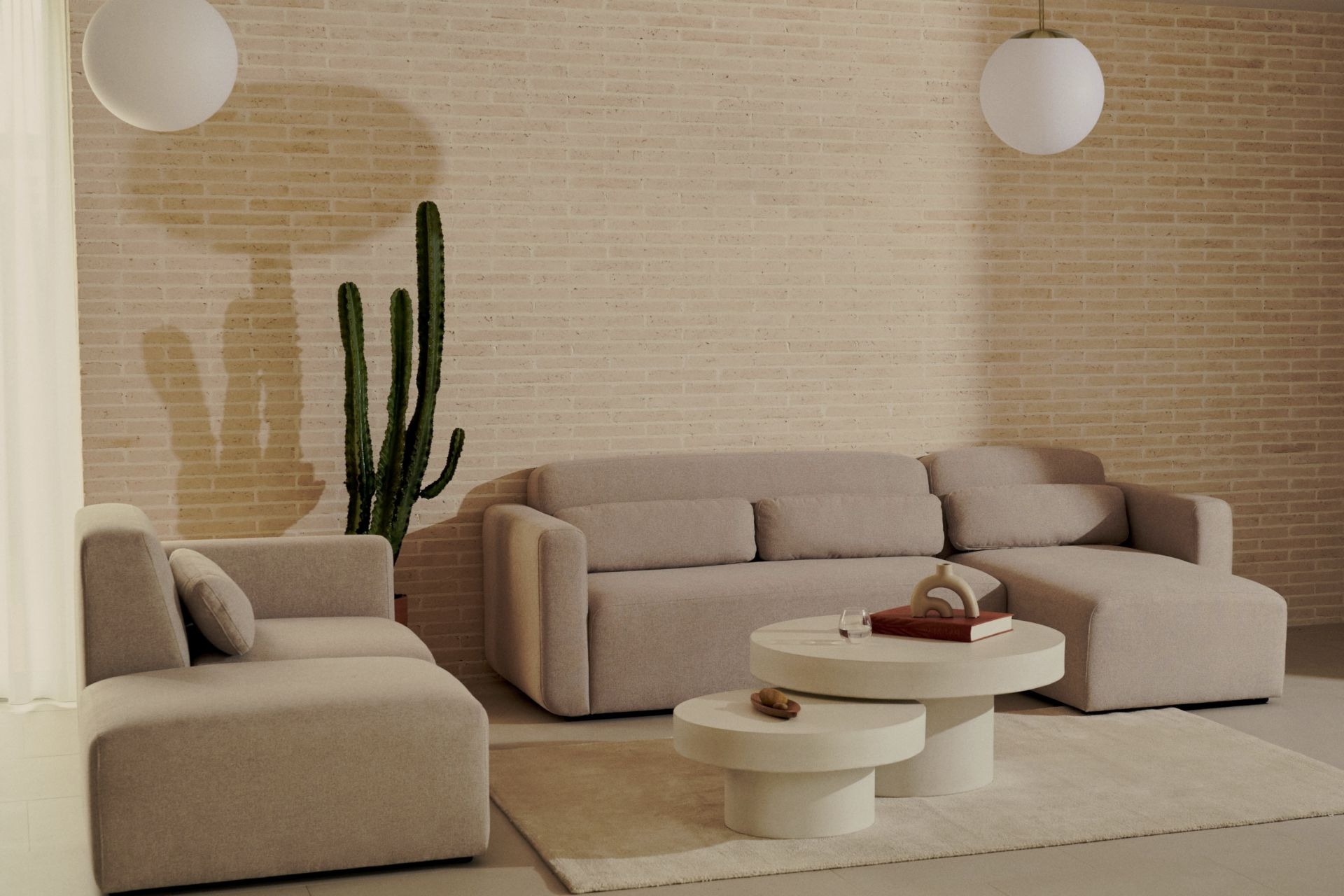 Hinten an einer hellen Ziegelwand steht ein beiges Sofaelement mit Stoffbezug, rechts daneben ein Weiteres, sodass sie in L-Form ausgerichtet sind. Vor ihnen liegt ein beiger flacher Teppich, auf dem ein 2-er Set runder cremefarbener Couchtische mit Sockelfuß steht. Rechts in der Ecke steht ein Kaktus und über den Sofaelementen hängt jeweils eine weiße Glaskugel-Pendellampe.