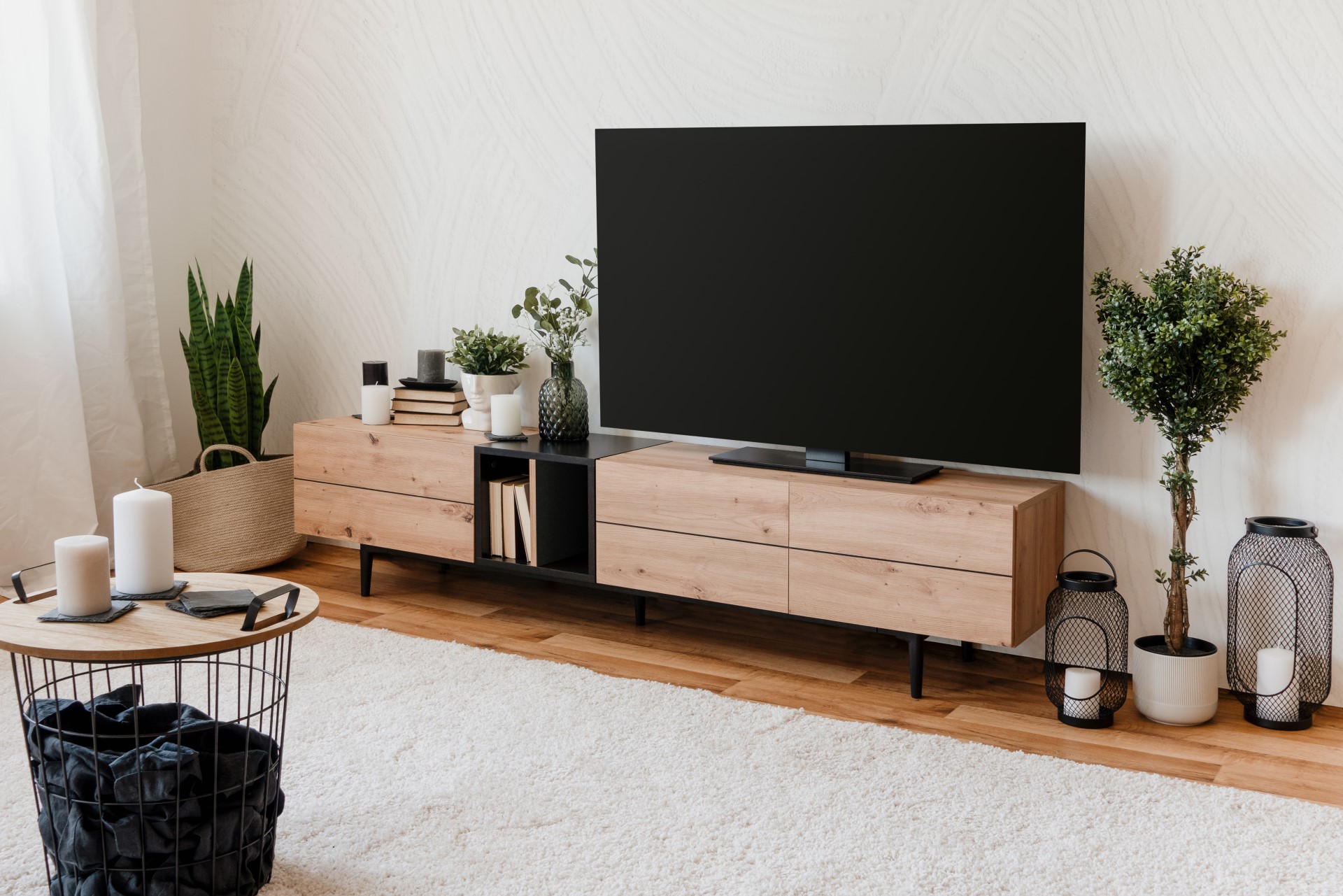 Modernes Wohnzimmer mit einem großen Fernseher auf einem Lowboard in Holzoptik, Pflanzen, Kerzen und Dekorationsartikeln.