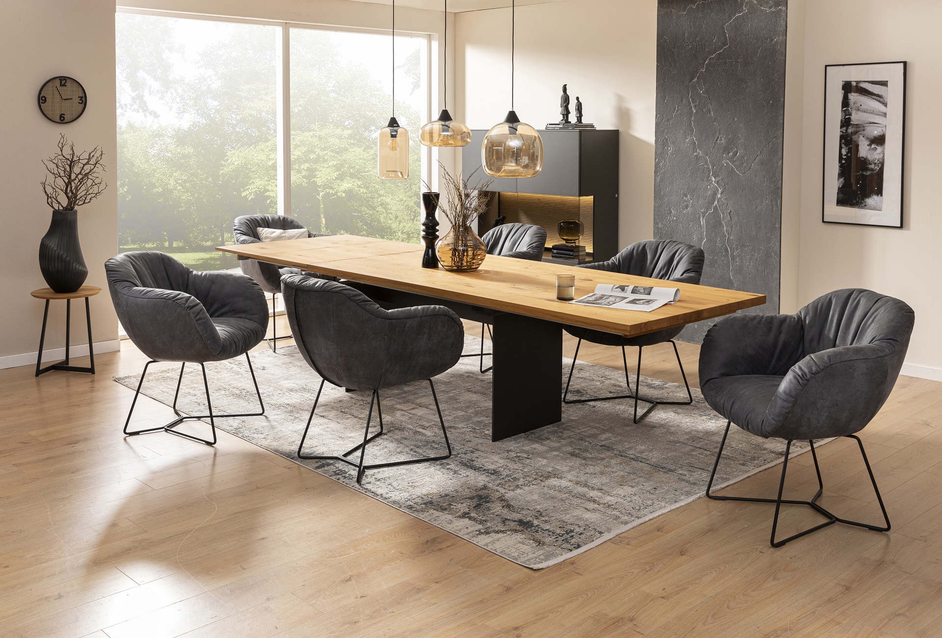 Modernes Esszimmer mit einem Tisch aus Holz und sechs grauen, gepolsterten Stühlen. Darüber hängt eine Pendelleuchte. Durch ein großes Fenster ist eine grüne Landschaft zu sehen.