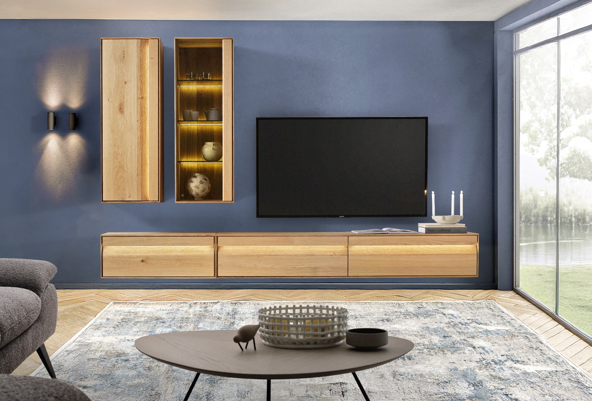 Moderne, grifflose Wohnwand der Marke Sormani aus hellem Holz und mit moderner Beleuchtung. Die Wand ist in einem intensiven Blauton gestrichen.