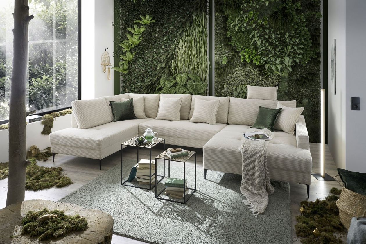 Auf dem Foto befindet sich eine cremefarbene Cord-Wohnlandschaft in U-Form mit einer dunkelgrünen bepflanzten Wand im Hintergrund und einem großen Fenster rechts. Das Sofa steht auf einem grauen Teppich hinter einem 2-er Satz Tische.