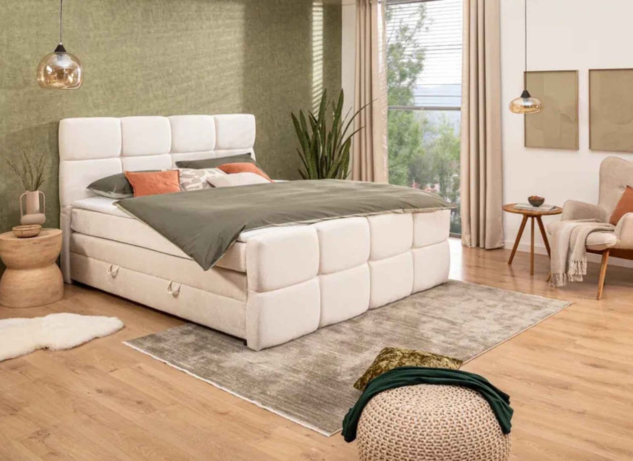 Modernes Schlafzimmer mit gepolstertem Bett, grüner Bettwäsche, Holzboden, Pflanzen und einem Sessel neben einem kleinen runden Tisch.