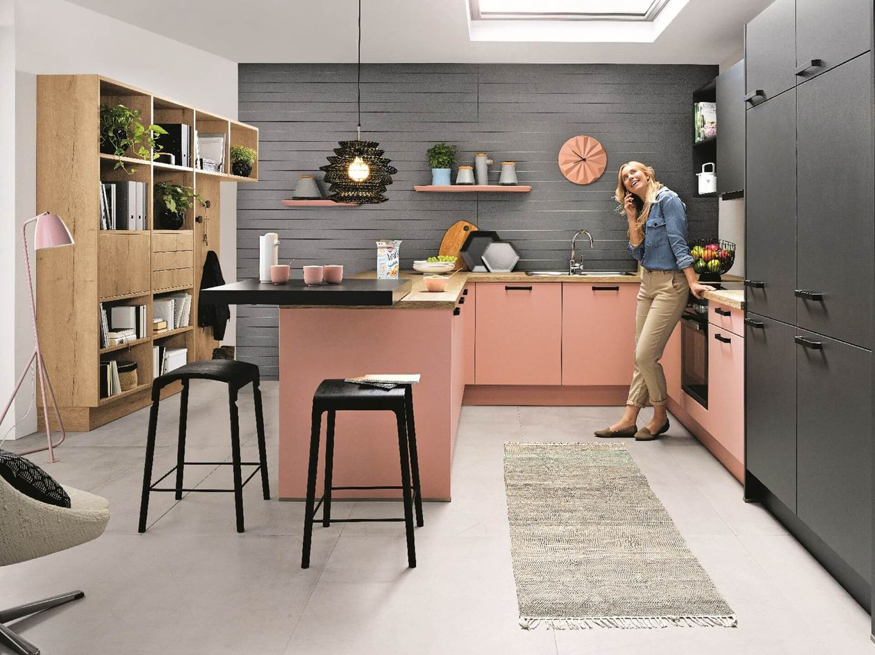 Moderne Küche mit rosa Schränken, schwarzen Arbeitsplatten und einer telefonierenden Frau. Offene Regale und Dekorationsgegenstände verschönern den Raum.