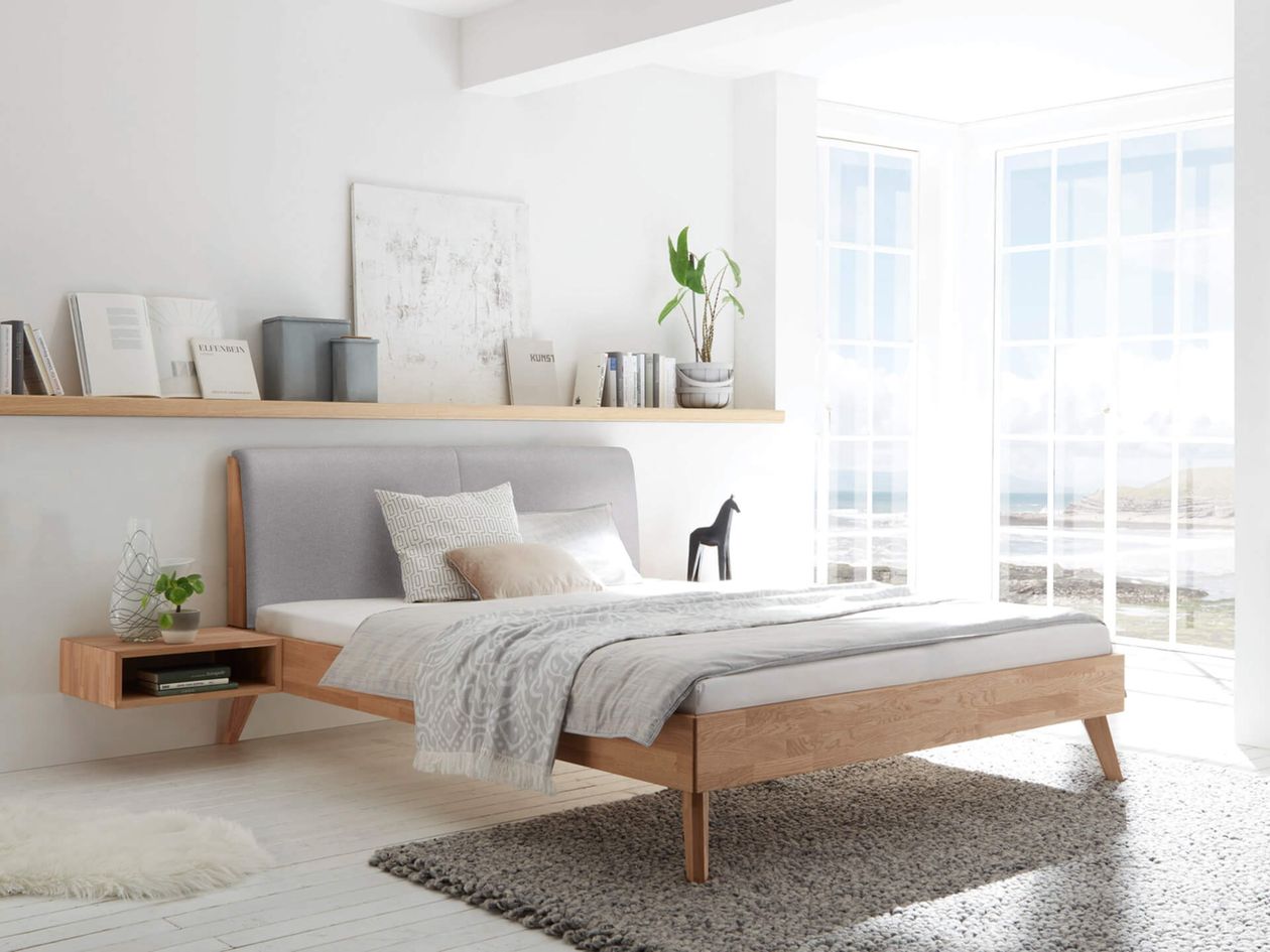 Minimalistisches Schlafzimmer mit einem Holzbett, grauem Kopfteil, gemütlicher Bettwäsche, schwebendem Nachttisch und einem großen Fenster mit malerischer Aussicht.