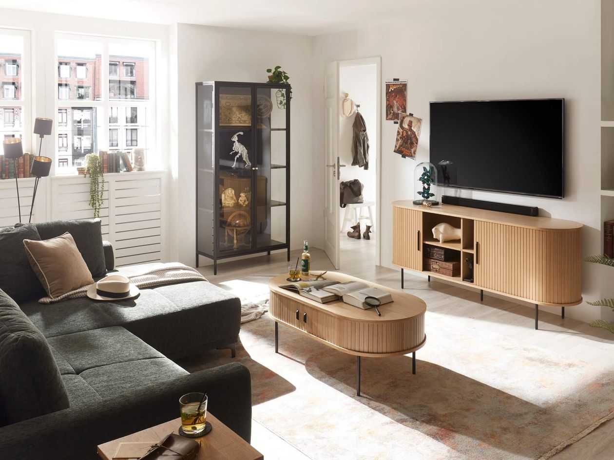 Beigefarbenes TV-Lowboard mit gerillter Front und abgerundeten Seiten in einem Wohnzimmer mit dunkelgrünem Sofa, Vitrine und Fernseher.
