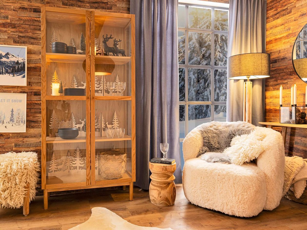 Gemütliches Wohnzimmer mit Holzschrank, Plüschsessel, Fellakzenten und Blick auf eine verschneite Landschaft durchs Fenster. Warmes Licht und rustikale Dekoration verstärken das Ambiente.