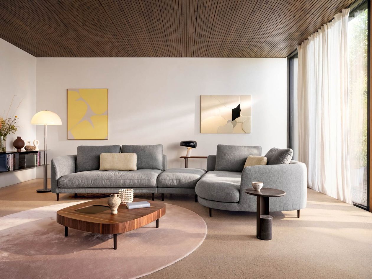 Elegantes graues Ecksofa mit organisch geschwungener Form in einem hellen Wohnzimmer mit Holzcouchtisch, rundem Teppich und modernen Kunstwerken an der Wand.