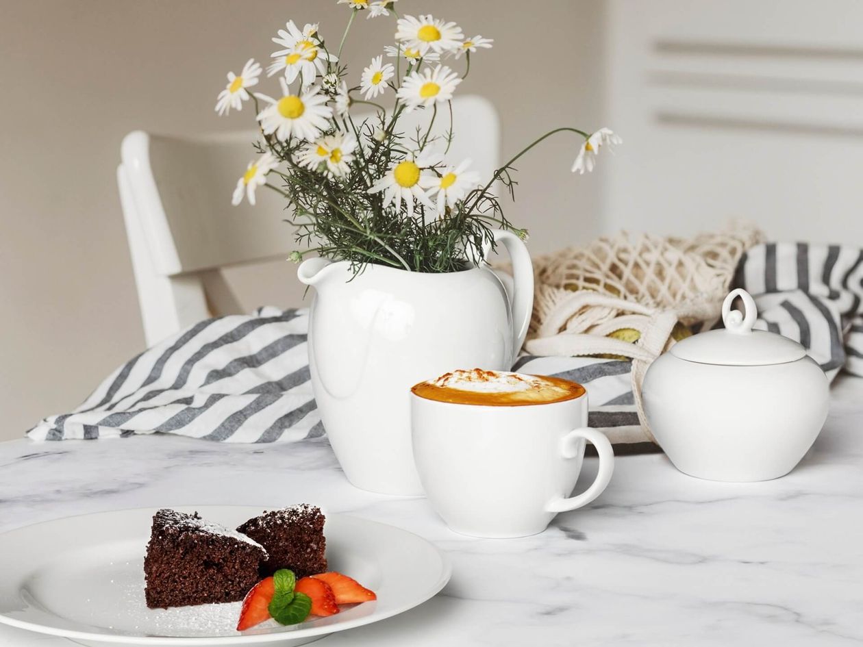 Weißer Frühstücksteller mit geschwungenem Rillenrand, einer Kaffeetasse und Zuckerdose auf einem Marmortisch, daneben ein Cappuccino, ein Stück Schokoladenkuchen mit Erdbeeren und ein Blumenstrauß aus Margeriten in einer weißen Kanne.