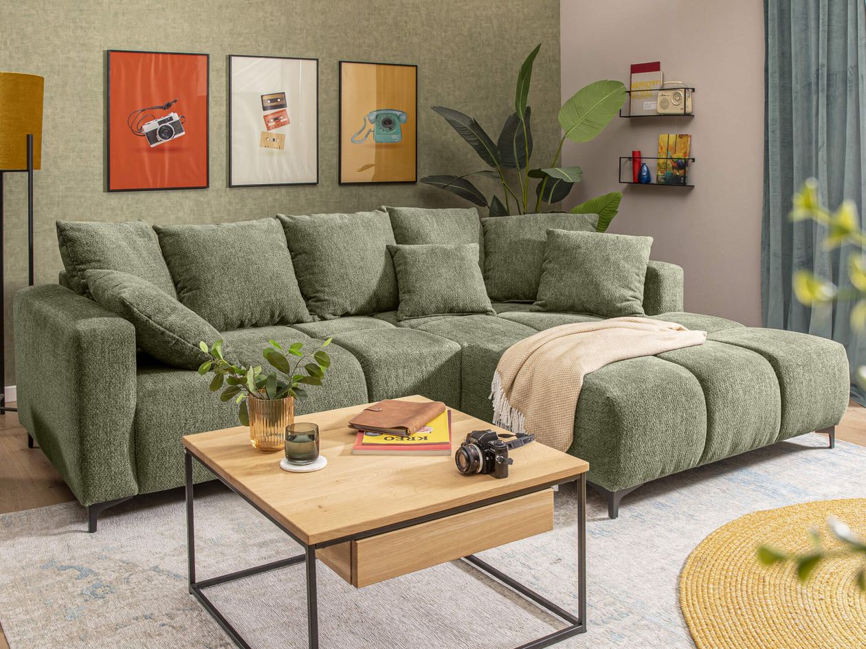 Grüne Eckcouch, Holztisch und bunte Wandbilder in hellem Wohnzimmer. Pflanzen und Stehlampe sorgen für Gemütlichkeit.