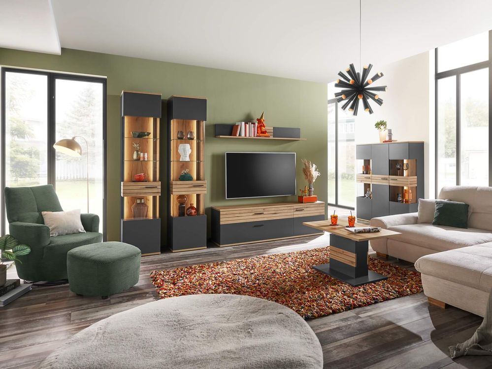 Modernes Wohnzimmer mit grünen Wänden, Fernseher, Regalen, grauen und beigen Sofas, rundem Teppich und geometrischem Kronleuchter. Große Fenster sorgen für natürliches Licht.