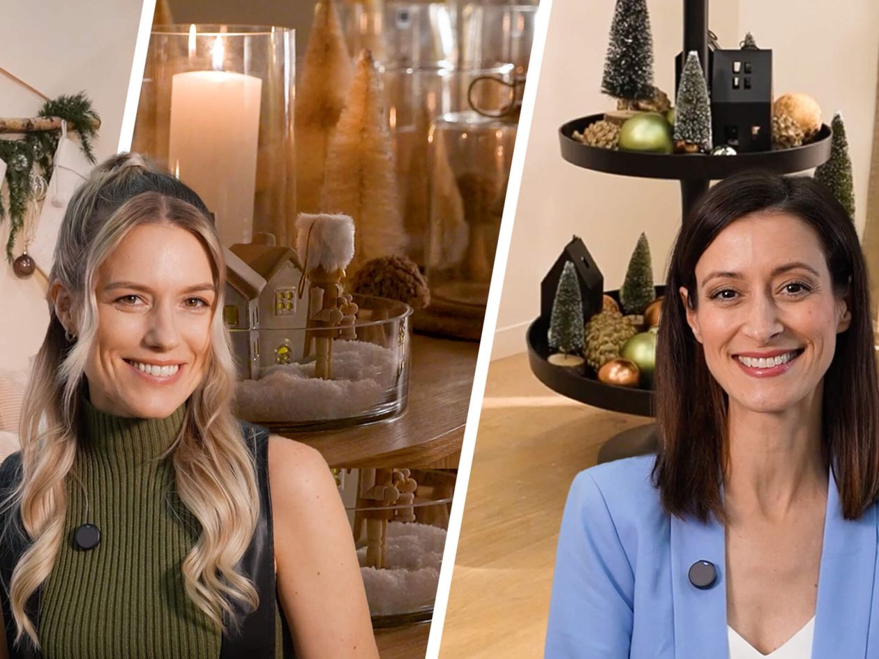 Festliche Dekoration mit Strümpfen, Kerzen und Lebkuchenhäusern. Zwei lächelnde Frauen, eine in Grün, die andere in Blau, mit mehrstöckiger Tablettdekoration.