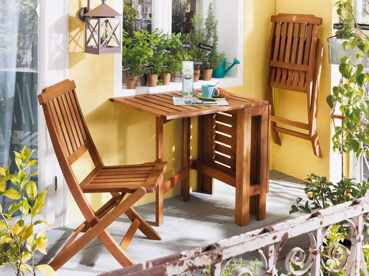 Gemütlicher Balkon mit einem Holztisch, zwei Stühlen, Topfpflanzen, einer Laterne und einer Tasse auf dem Tisch vor einer gelben Wand.
