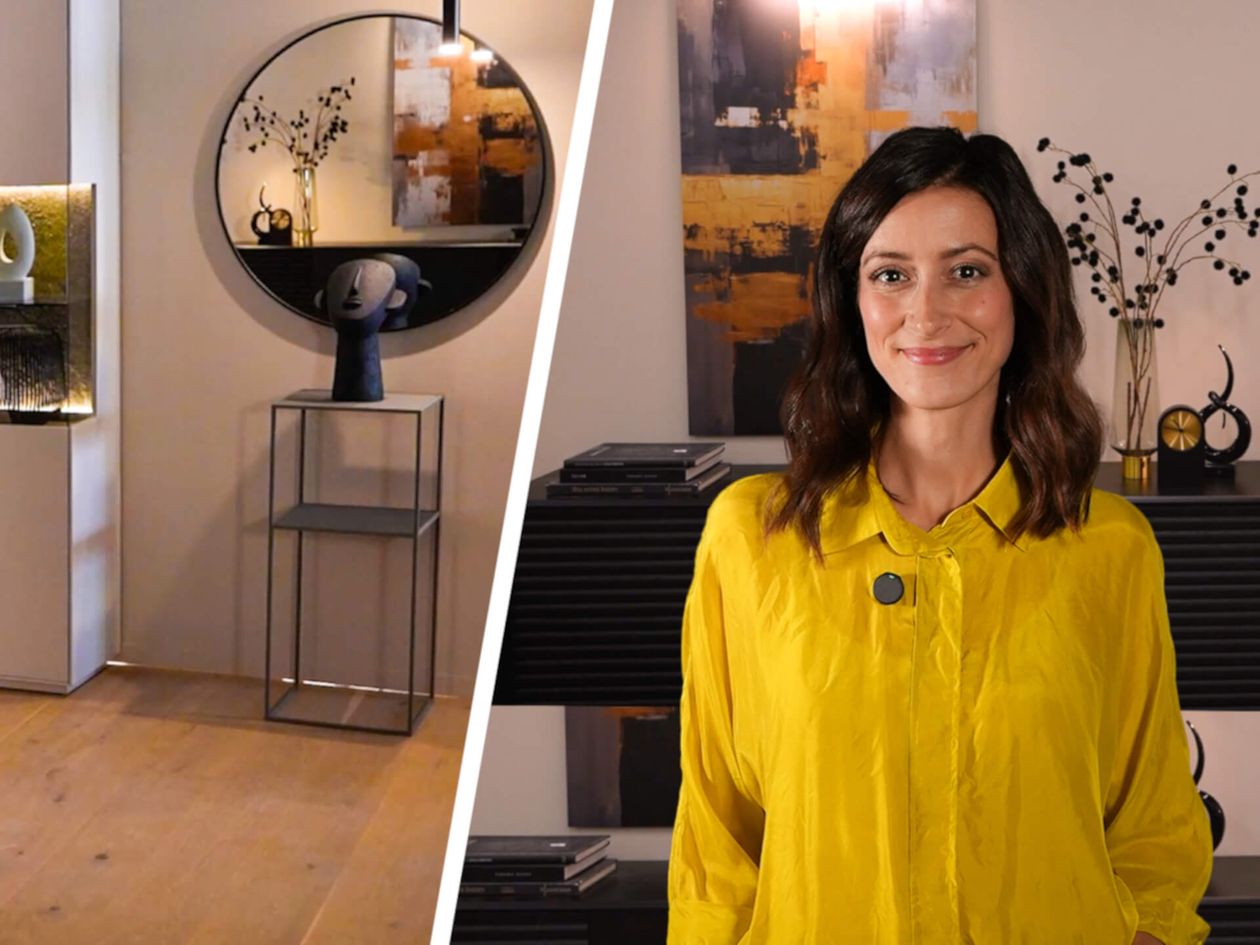 Eine Frau in gelber Bluse lächelt in einem modern eingerichteten Raum mit Kunstwerken und Spiegeln.
