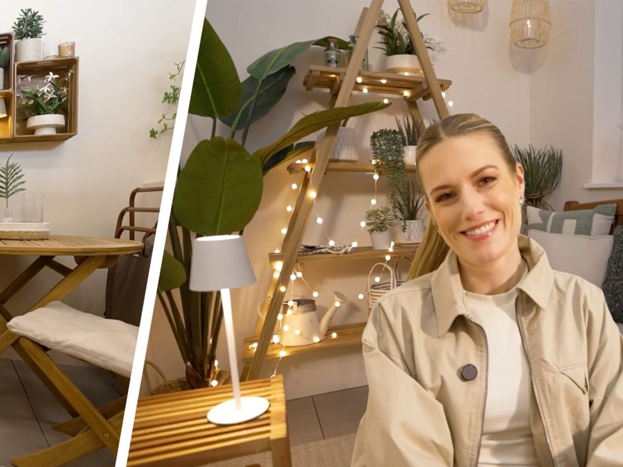 Eine lächelnde Frau sitzt in einem gemütlich eingerichteten Raum mit Pflanzen, einem Holzregal in Form einer Leiter mit Lichtern sowie einem kleinen Tisch und einem Stuhl aus Holz.