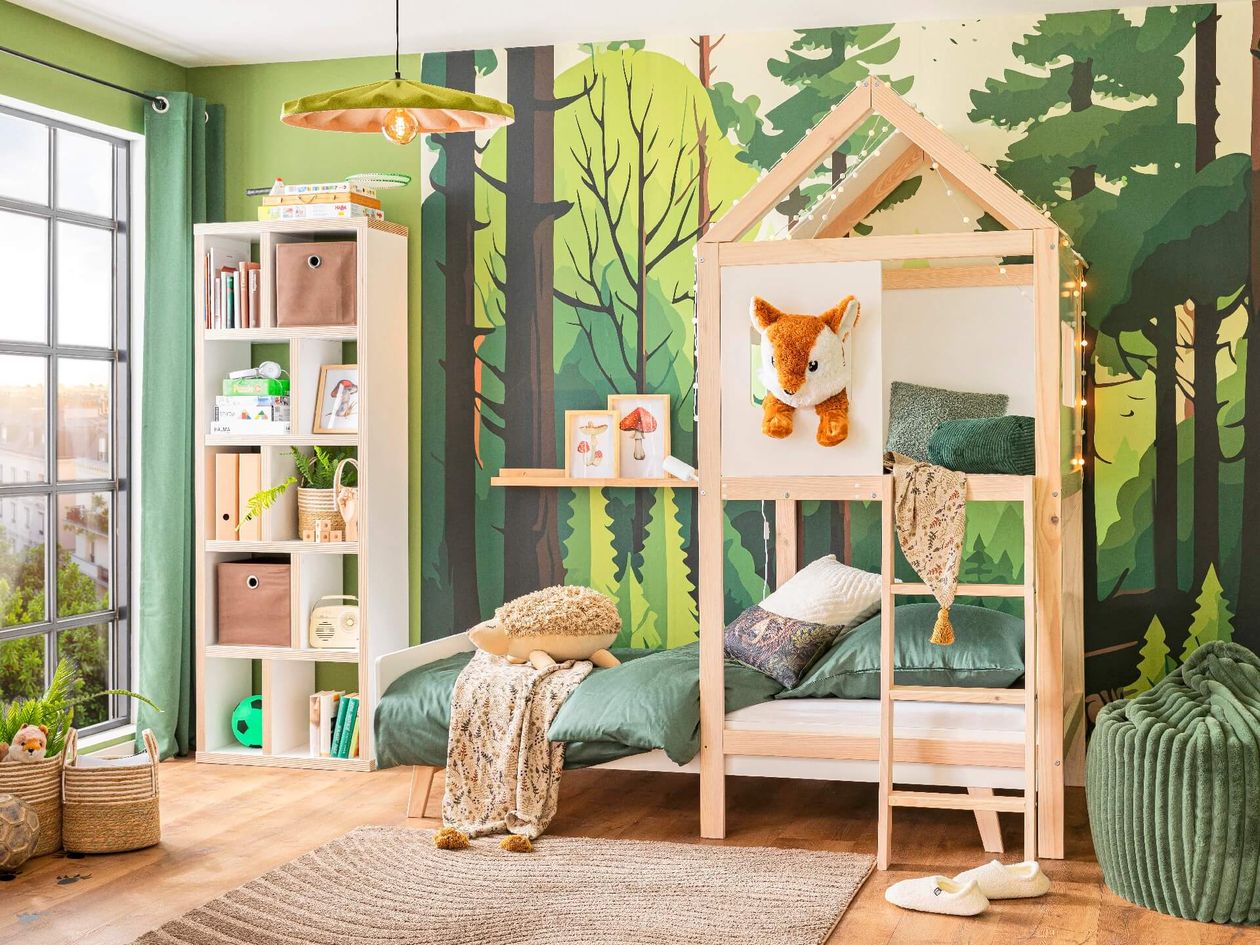 Kinderzimmer mit einer Tapete zum Thema Wald, einem hölzernen Bett in Form eines Hauses, grüner Bettwäsche, Spielzeug, Regalen mit Büchern und einem großen Fenster.