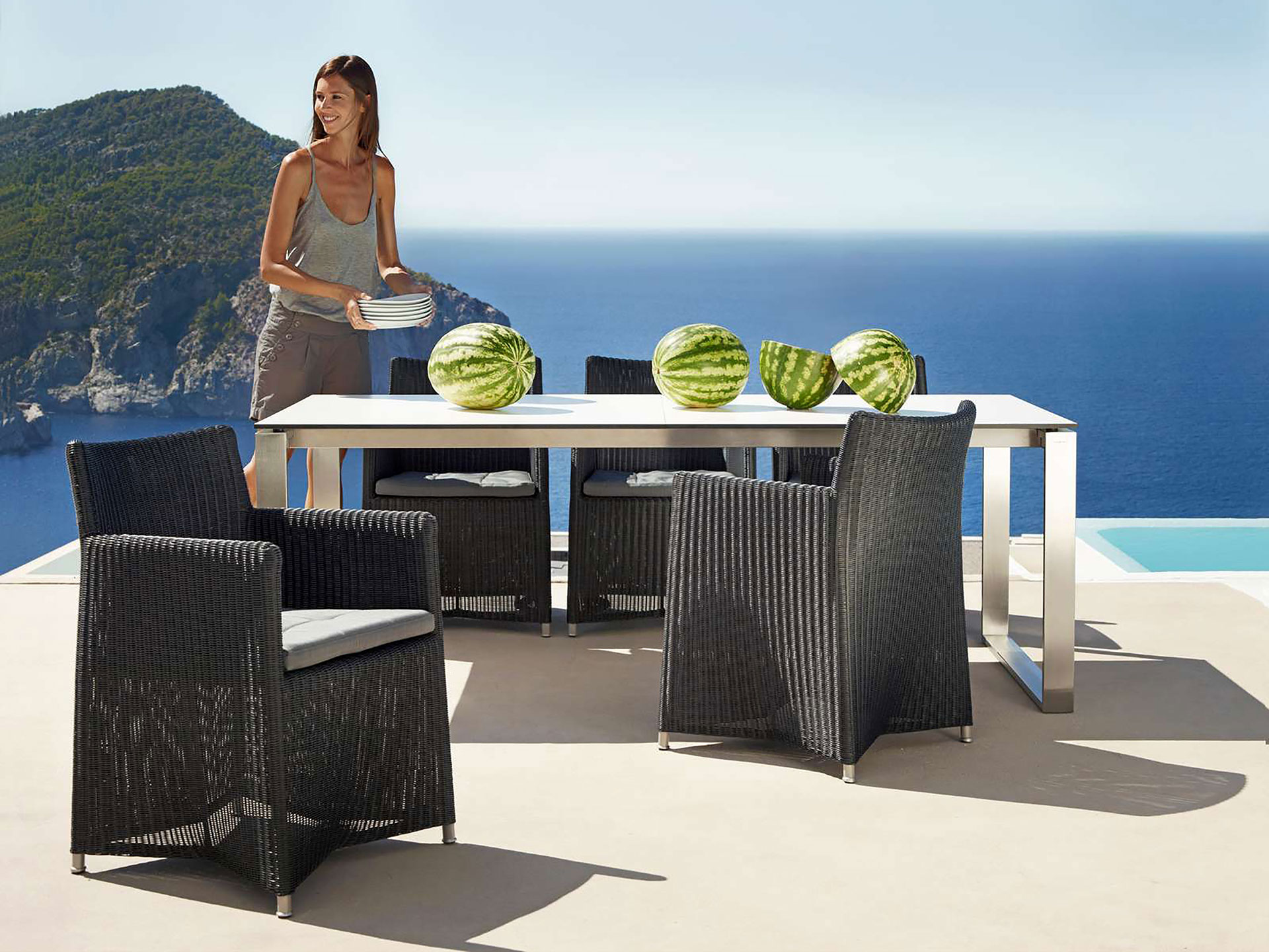Mehrere Gartensessel des Modells Diamond von Cane-line mit Geflecht in Graphit stehen um einen  Metallgartentisch mit großen Wassermelonen. Eine Frau trägt Teller herbei. Im Hintergrund ist eine Insel im Meer zu sehen.