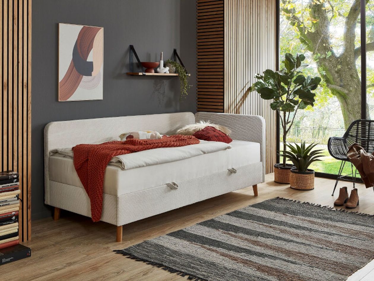 Modernes, helles Schlafzimmer mit kleinem Bett, auf dem eine rotte Strickdecke liegt. Große Zimmerpflanzen sorgen für eine natürliche Atmosphäre.