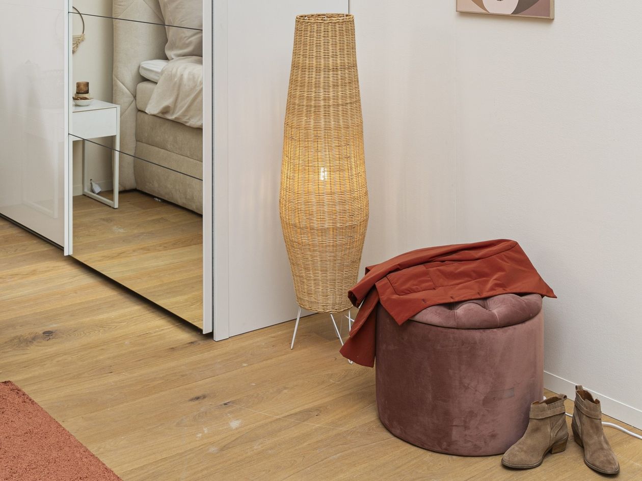 Stehlampe Kamaria der Marke Daheim aus braunem Rattan neben einem Schrank stehend